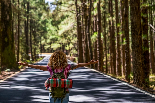 Rückansicht der lockigen Frau, die die Arme öffnet und die Straßenfahrt mit Rucksack genießt - alternativer Menschenfreiheitslebensstil, der auf der Straße mitten in einem hohen Baumwald geht