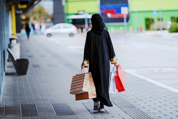 Rückansicht der muslimischen Frau in traditioneller Kleidung, die draußen mit Einkaufstüten in Händen geht. Mode ist für alle. Diversity-Konzept.