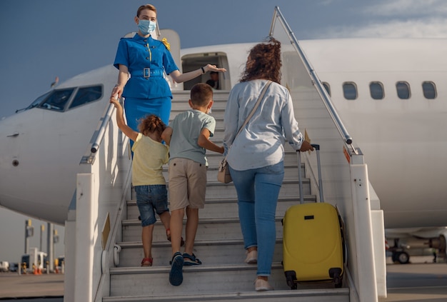 Rückansicht der Mutter mit zwei kleinen Kindern und Koffer beim Einsteigen in das Flugzeug. Flugstewardess mit Schutzmaske begrüßt die Familie. Menschen, Reisen, Urlaubskonzept