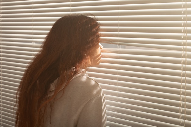 Rückansicht der schönen jungen Frau mit dem langen Haar, das durch Jalousien späht