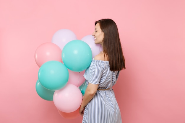 Rückansicht der schönen zarten jungen glücklichen Frau im blauen Kleid halten bunte Luftballons, die einzeln auf hellrosa Hintergrund beiseite schauen. Geburtstagsfeier, Konzept der aufrichtigen Emotionen der Menschen.