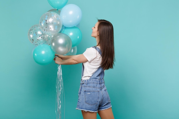 Rückansicht der süßen jungen brünetten Frau in Denim-Kleidung, die bunte Luftballons einzeln auf türkisfarbenem Wandhintergrund feiert und hält. Geburtstagsfeier, Menschen Emotionen Konzept.