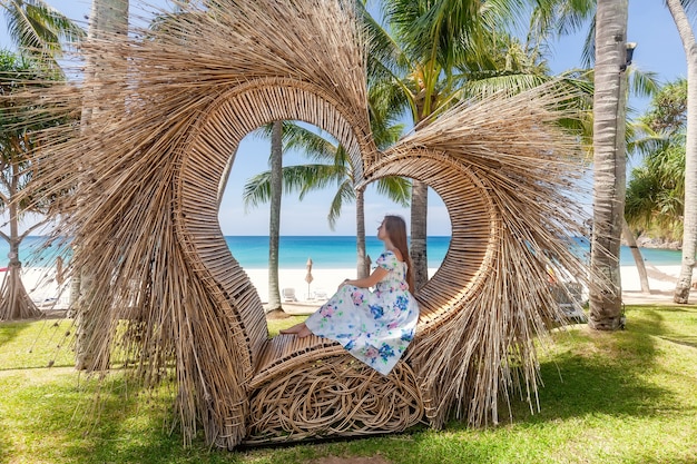 Rückansicht der Touristenfrau, die im Foto-Spot sitzt, wie Strohherz mit tropischen Palmen und blauem Meer mit weißem Sandstrand im Hintergrund. Natur- und Reisekonzept