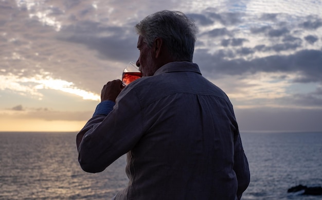 Rückansicht des älteren Mannes, der bei Sonnenuntergang vor dem Meer sitzt und ein Glas Wein trinkt Horizont über dem Wasser Einsamer alter Mann ertränkt seine Probleme in Alkohol