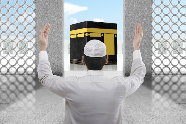 Foto rückansicht des asiatischen muslimischen mannes, der mit kaaba-ansicht und blauem himmelshintergrund steht und betet