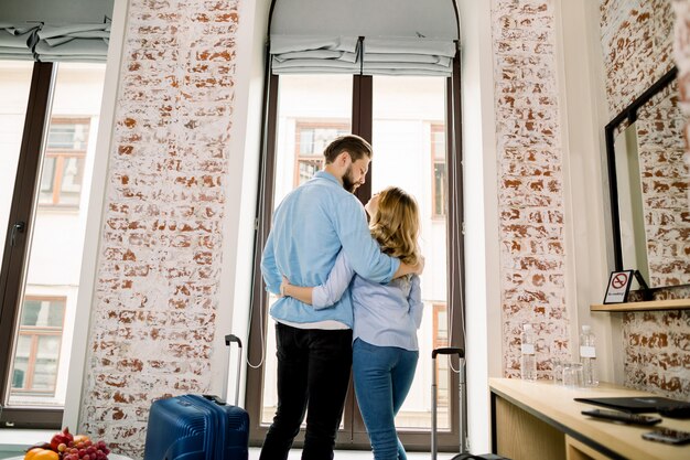 Rückansicht des glücklichen liebenden Paares im Hotelzimmer, das das Fenster umarmt und steht