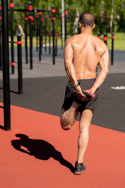 Rückansicht des hemdlosen jungen Sportlers, der seinen linken Fuß in der Hand hält, während er sich vor dem Training auf Sportanlagen aufwärmt