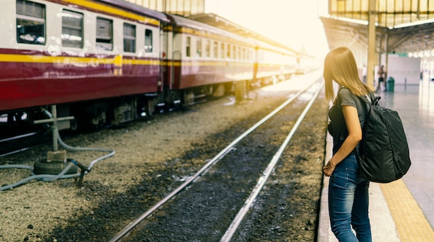 Rückansicht des jungen asiatischen weiblichen Reisenden mit kleinem Rucksack auf der Eisenbahnstation. Warten auf Zug mit Transport auf Urlaubsreise. Reisekonzept