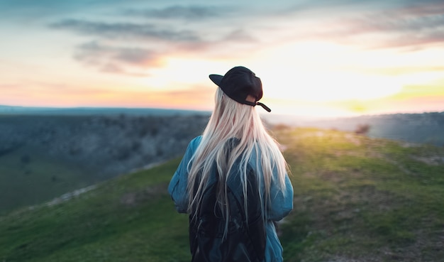 Rückansicht des jungen blonden Mädchens mit der schwarzen Kappe und dem Rucksack, die auf Spitze der Hügel gehen. Hintergrund des Sonnenuntergangs.