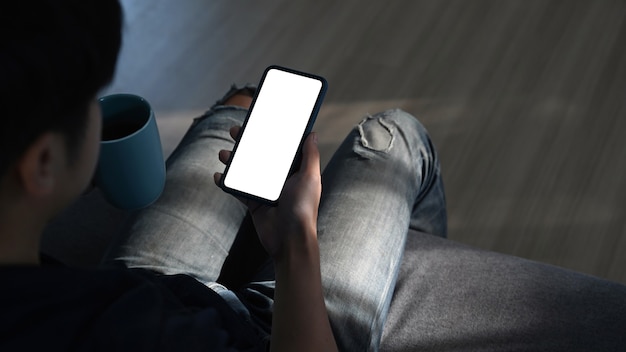 Rückansicht des jungen Mannes, der Smartphone benutzt und eine Tasse Kaffee hält, während er auf dem Sofa sitzt.