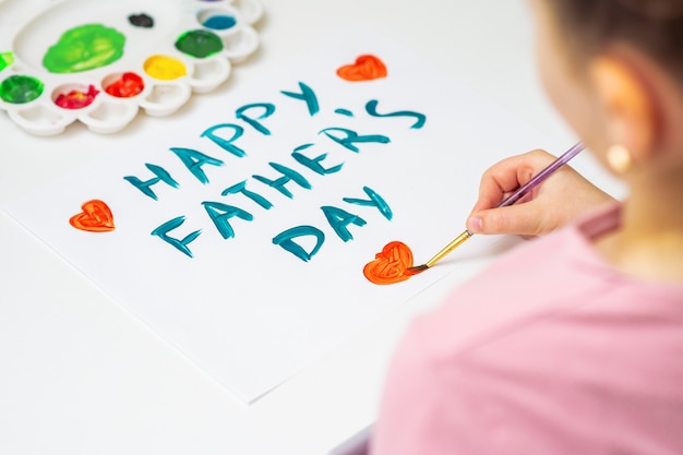 Rückansicht des kleinen Mädchens zeichnet rotes Herz mit Text Happy Father's Day Grußkarte von Aquarellen auf weißem Papier. Konzept zum Vatertag.