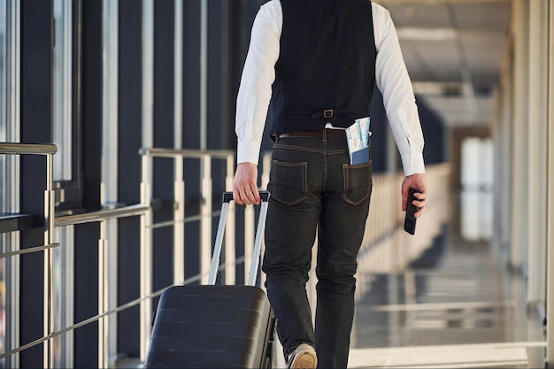 Rückansicht des männlichen Passagiers in eleganter formeller Kleidung befindet sich in der Flughafenhalle mit Tickets in der Tasche.