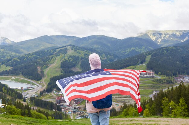 Rückansicht des schlanken aktiven Touristen, der Freizeitaktivitäten liebt, wandern, US-Flagge halten, ihren Rücken damit bedecken, Berge betrachten