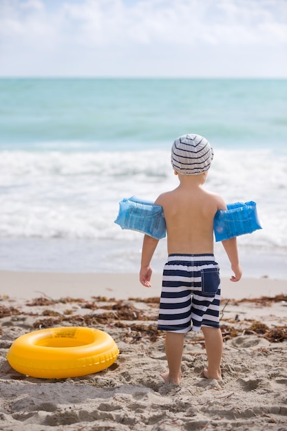 Rückansicht des süßen Kleinkindjungen mit aufblasbaren Sicherheitsarmbändern, die am Strand stehen. Sicherheit zuerst