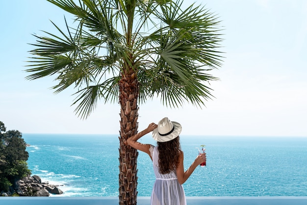 Rückansicht: Ein Mädchen in Hut und weißem Kleid bewundert den Blick auf das Meer in der Nähe einer Palme am Rand des Pools. Spa-Geschichten