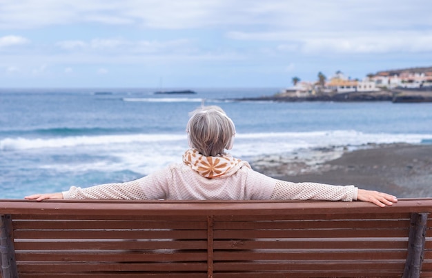 Rückansicht einer älteren Frau mit Kopfhörern, die im Freien auf dem Meer sitzt und den Horizont genießt und die Freiheit und den Urlaub genießt Kaukasische ältere Frau, die sich an einem sonnigen Tag in der Nähe des Strandes entspannt