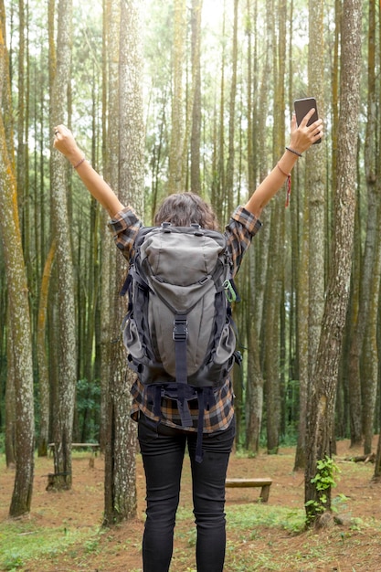 Rückansicht einer asiatischen Frau, die im Wald wandert und ein Mobiltelefon mit aufgeregtem Gesichtsausdruck in der Hand hält