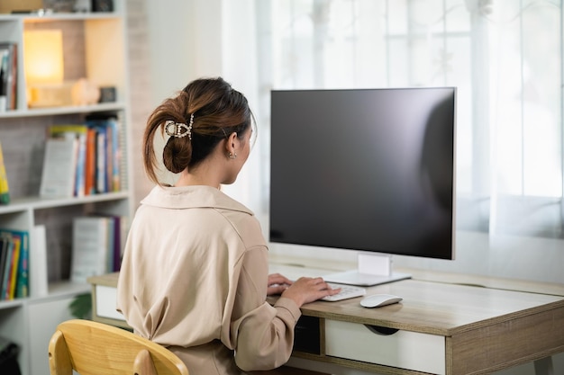 Rückansicht einer asiatischen freiberuflichen Frau, die zu Hause auf der Tastatur tippt und am Laptop auf einem Holztisch arbeitet Unternehmerin, die für ihr Unternehmen im Wohnzimmer zu Hause arbeitet Business Work Home-Konzept