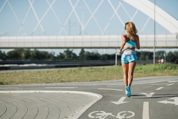 Rückansicht einer erwachsenen Lauffrau, die auf der Stadtpromenade am Flussufer joggt und Musik hört.