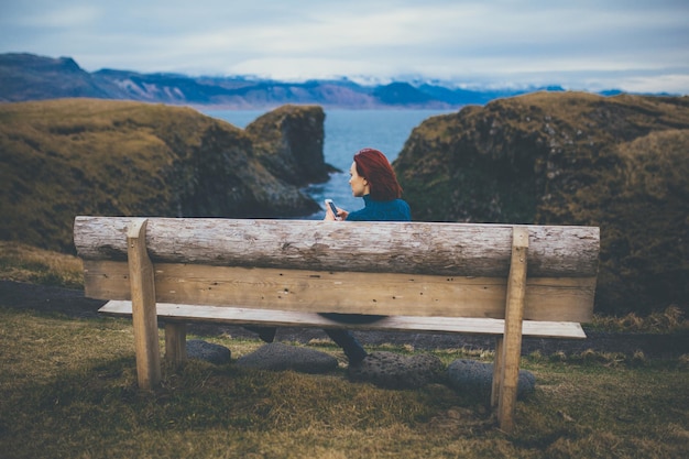 Rückansicht einer Frau, die auf einer Bank sitzt und ein Smartphone in Island benutzt