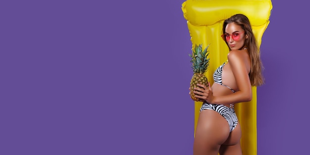 Rückansicht einer Frau, die im Bikini mit gelber aufblasbarer Matratze posiert, während sie Ananas hält