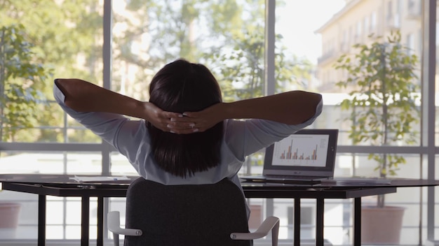 Rückansicht einer Frau, die im Büro am Computer arbeitet und sich am Schreibtisch streckt