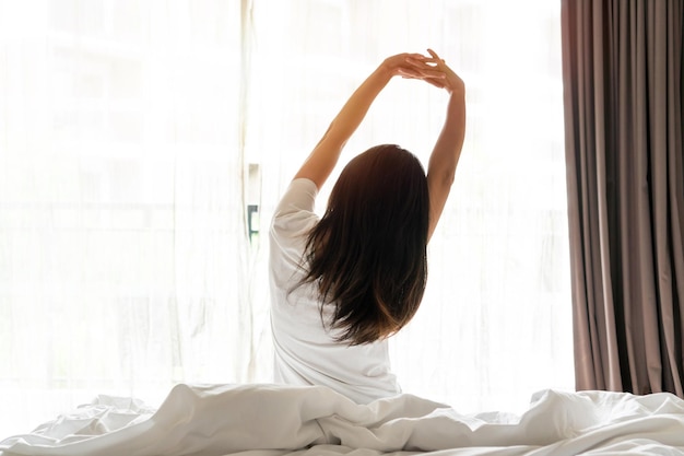 Rückansicht einer Frau, die sich nach dem Aufwachen mit Sonnenlicht morgens im Schlafzimmer zu Hause streckt