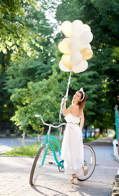 Rückansicht einer Frau, die über ihre Schulter in die Kamera lächelt und mit einem Fahrrad im Park posiert, der farbige Luftballons hält