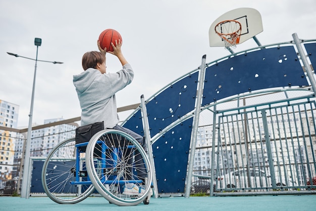 Rückansicht einer Frau mit Behinderung, die im Rollstuhl sitzt und den Ball im Freien in den Korb wirft