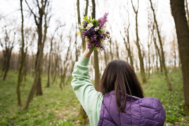 Rückansicht einer Frau mit erhobener Hand, die einen Frühlingsblumenstrauß im Wald hält. Outdoor-Freizeitkonzept