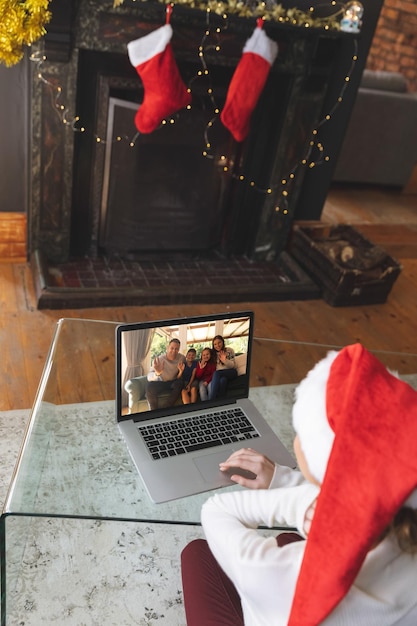 Rückansicht einer Frau mit Weihnachtsmannmütze, die zu Hause einen Videoanruf mit ihrer Familie in Weihnachtsmannmützen führt und auf dem Laptop winkt. soziale distanzierung während des quarantäne-sperrkonzepts für das weihnachtsfest.