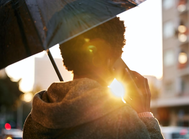 Rückansicht einer Geschäftsfrau, die einen Regenschirm und ein Smartphone benutzt, während sie im Regen vor einem städtischen Hintergrund spazieren geht