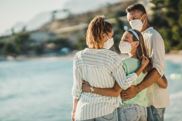 Rückansicht einer glücklich umarmten Familie mit schützender N95-Maske, die bei einer Koronapandemie Zeit am Strand verbringt.