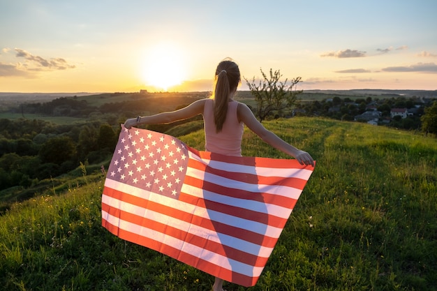 Rückansicht einer glücklichen jungen Frau, die bei Sonnenuntergang mit der USA-Nationalflagge im Freien posiert. Positives Mädchen, das den Unabhängigkeitstag der Vereinigten Staaten feiert. Internationaler Tag des Demokratiekonzepts.