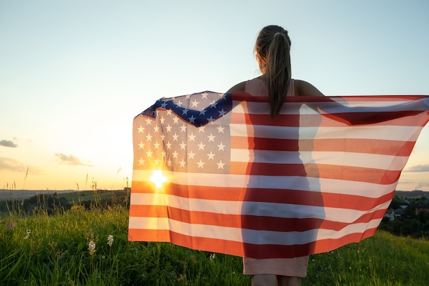 Rückansicht einer glücklichen jungen Frau, die mit der Nationalflagge der USA bei Sonnenuntergang im Freien posiert. Positives Mädchen, das den Unabhängigkeitstag der Vereinigten Staaten feiert. Internationaler Tag des Demokratiekonzepts.