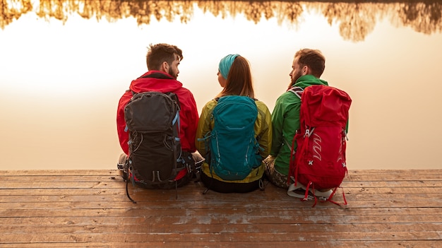 Rückansicht einer Gruppe junger Wanderer in bunter warmer Oberbekleidung und mit Rucksäcken, die auf einem hölzernen Pier in der Nähe des Sees sitzen und sprechen, während sie sich nach dem gemeinsamen Trekking in der Herbstnatur ausruhen