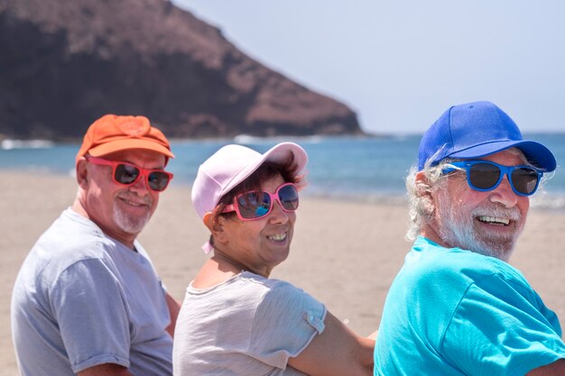 Rückansicht einer Gruppe von drei Freunden, die am Strand sitzen, erwachsene Menschen im Ruhestand im Urlaub im Freien auf See lächeln und genießen Sonne und Sommer