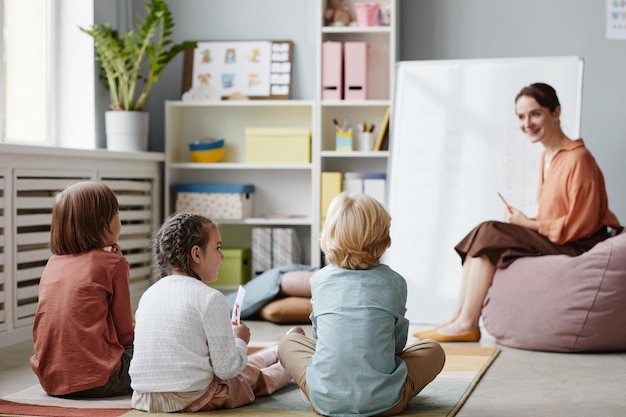 Rückansicht einer Gruppe von Kindern, die auf dem Boden sitzt und dem Lehrer während des Unterrichts im Klassenzimmer zuhört