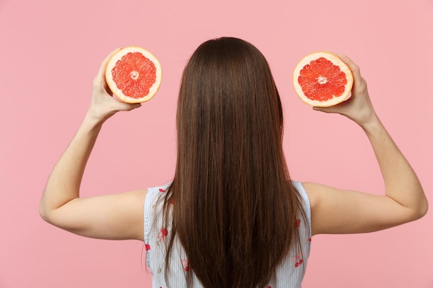 Rückansicht einer jungen Frau in Sommerkleidung, die Hälften frischer, reifer Grapefruit in den Händen hält, isoliert auf rosafarbenem Pastellwandhintergrund. Menschen lebendiger Lebensstil entspannen Urlaubskonzept. Mock-up-Kopienbereich.