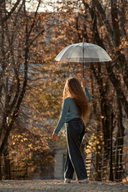 Rückansicht einer jungen Frau mit langen Haaren, die durch den Herbstpark spaziert Hübsches Mädchen mit transparentem Regenschirm