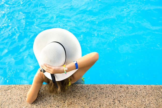 Rückansicht einer jungen Frau mit langen Haaren, die einen gelben Strohhut trägt und sich an einem sonnigen Tag im warmen Sommerpool mit blauem Wasser entspannt.