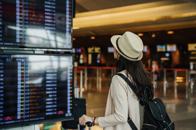 Rückansicht einer jungen reisenden Touristenfrau mit Hut, die auf den Fahrplan blickt und in der Lobbyhalle am internationalen Flughafen wartet. weibliche Passagiere reisen am Wochenende ins Ausland. Flugkonzept