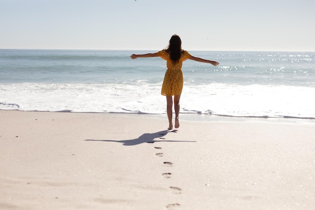 Rückansicht einer kaukasischen Frau mit langen dunklen Haaren, die ein gelbes Sommerkleid trägt und barfuß an einem idyllischen, sonnigen Strand läuft, die Arme in die Luft gestreckt, mit blauem Himmel und Meer im Hintergrund