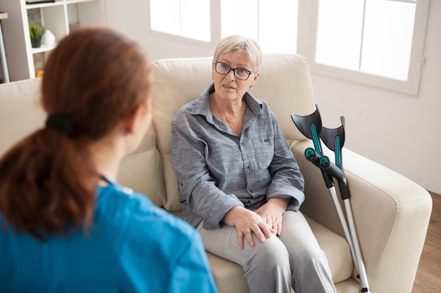 Rückansicht einer Krankenschwester in einem Pflegeheim, die mit einer Rentnerin spricht, die mit Krücken neben ihr auf der Couch sitzt.