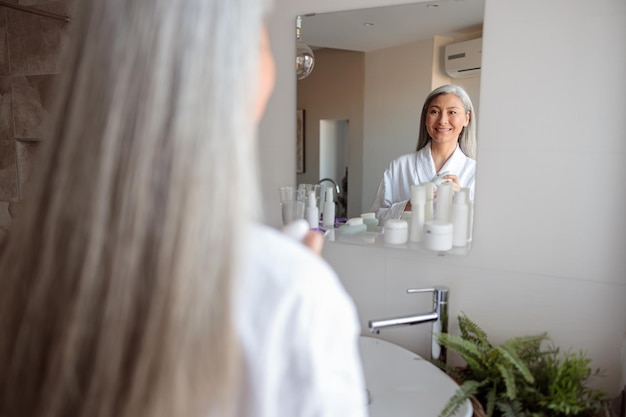 Rückansicht einer lächelnden Dame, die im Badezimmer in den Spiegel schaut, in einem weißen Bademantel gekleidet ist und eine Kosmetikflasche in den Händen hält