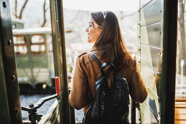 Rückansicht einer lächelnden jungen Frau mit einem Rucksack auf dem Rücken, die in einem Abenteuer mit dem Retro-Zug unterwegs ist.