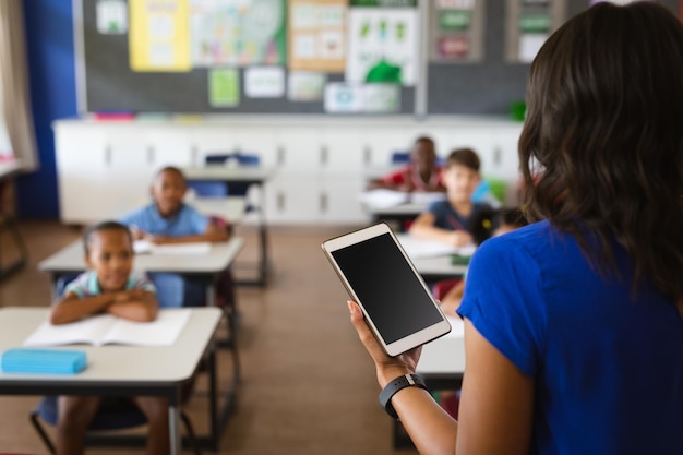 Rückansicht einer Lehrerin mit digitalem Tablet, die Schüler in der Klasse in der Schule unterrichtet