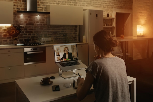 Rückansicht einer Mitarbeiterin, die aus der Ferne arbeitet und die Rede eines Chefs während einer Videokonferenz auf einem Laptop von zu Hause aus notiert. Ein Business-Team bei einem Online-Meeting am Abend.