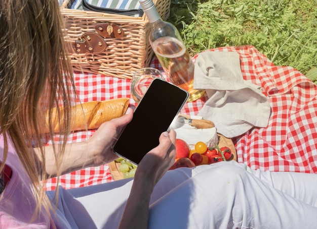 Rückansicht einer nicht erkennbaren Frau in weißen Hosen, die draußen picknickt und mit dem Smartphone fotografiert