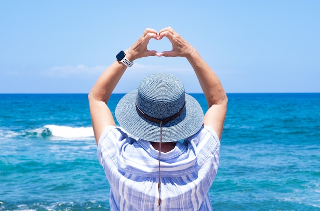 Rückansicht einer reifen Frau mit blauem Hut mit Blick auf den Horizont über dem Wasser, die Herzform mit den Händen macht. Konzept der Liebe und Freiheit